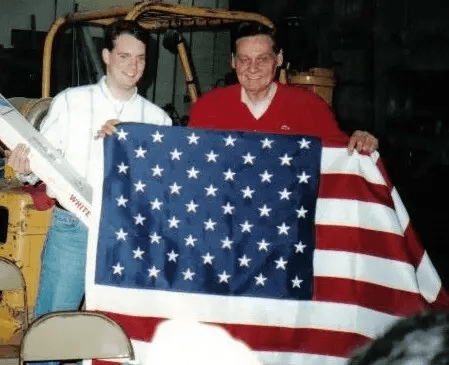 Two men holding up American Flag in front of warehouse vehicle