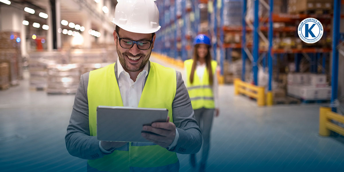 A smiling man in safety gear looks at an ipad in a warehouse.