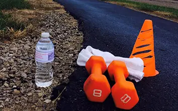 A water bottle, two dumbbell weights, a cone, and a towel placed on the side of a road